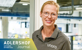 Lächelnde Frau mit Brille und grauem Poloshirt mit Aufschrift 'unora' vor hellem, industriellem Hintergrund