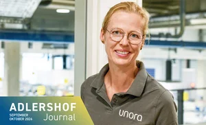 Lächelnde Frau mit Brille und grauem Poloshirt mit Aufschrift 'unora' vor hellem, industriellem Hintergrund
