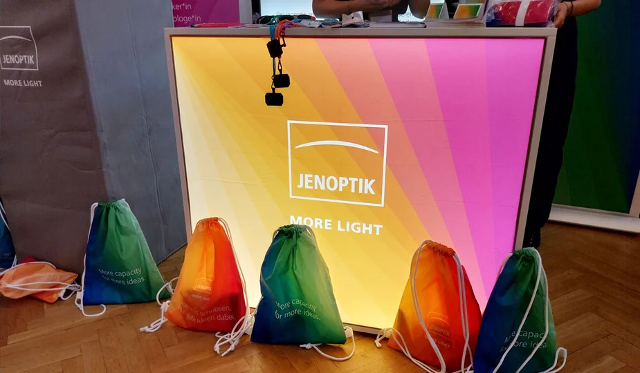 Leuchtender Messetresen mit dem Schriftzug 'JENOPTIK MORE LIGHT' und fünf bunte Stoffbeutel davor auf Holzboden