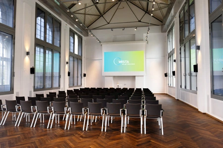 Hans-Grade-Saal mit Reihen von Stühlen vor einer Leinwand und hohen Fenstern an den Seiten