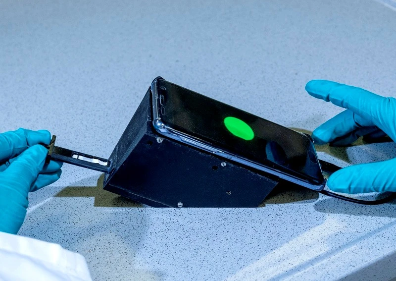 Schwarzer Kasten mit einem Teststreifen, darauf liegt ein Smartphone, das grünes Licht anzeigt