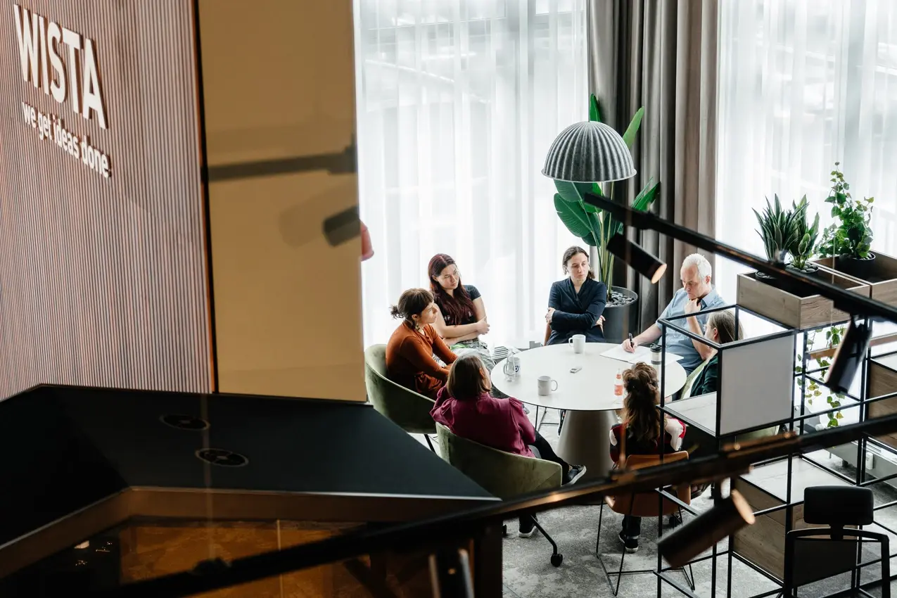 Round table by a window seat where seven people sit and talk together.