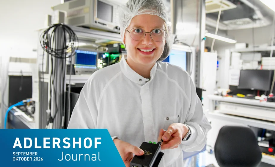 Lächelnde Person mit Schutzhaube, Brille und weißem Kittel hält ein elektronisches Gerät in einem Labor oder einer Produktionsumgebung