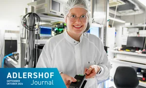 Lächelnde Person mit Schutzhaube, Brille und weißem Kittel hält ein elektronisches Gerät in einem Labor oder einer Produktionsumgebung