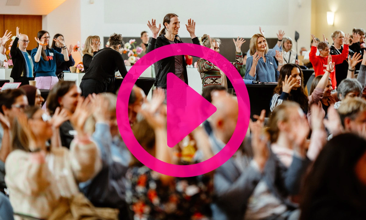 Gruppe von Menschen in einem Saal mit erhobenen Händen und einem großen pinkfarbenen Play-Symbol in der Bildmitte